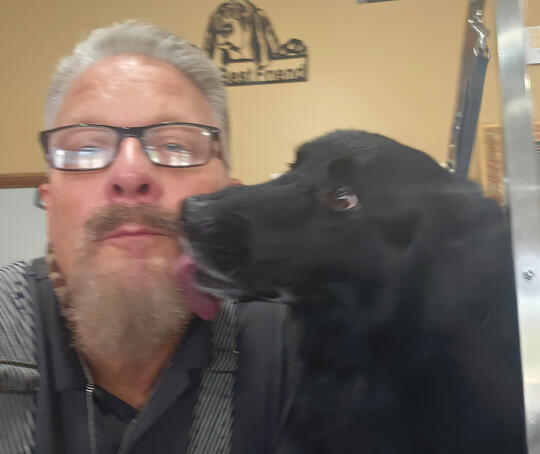 A profile picture of a man with mid-length gray hair, a gray beard, and mustache smiling inside a dog grooming salon. An American flag and framed artworks of dogs are seen in the background.